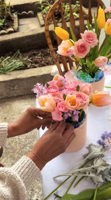Hands arranging peach roses, tulips and dusty miller into a floral box at the worktable