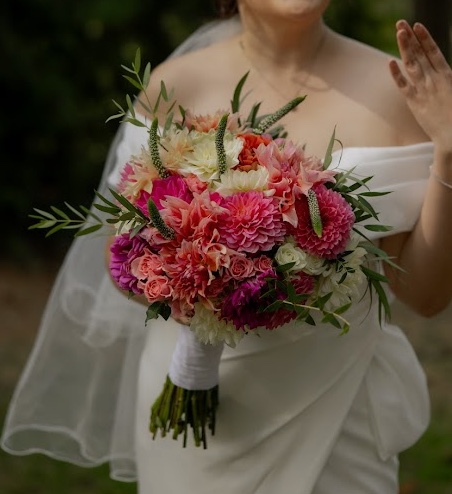 A bride in a white dress and veil holding a hand-tied bouquet of dahlias in pinks and corals