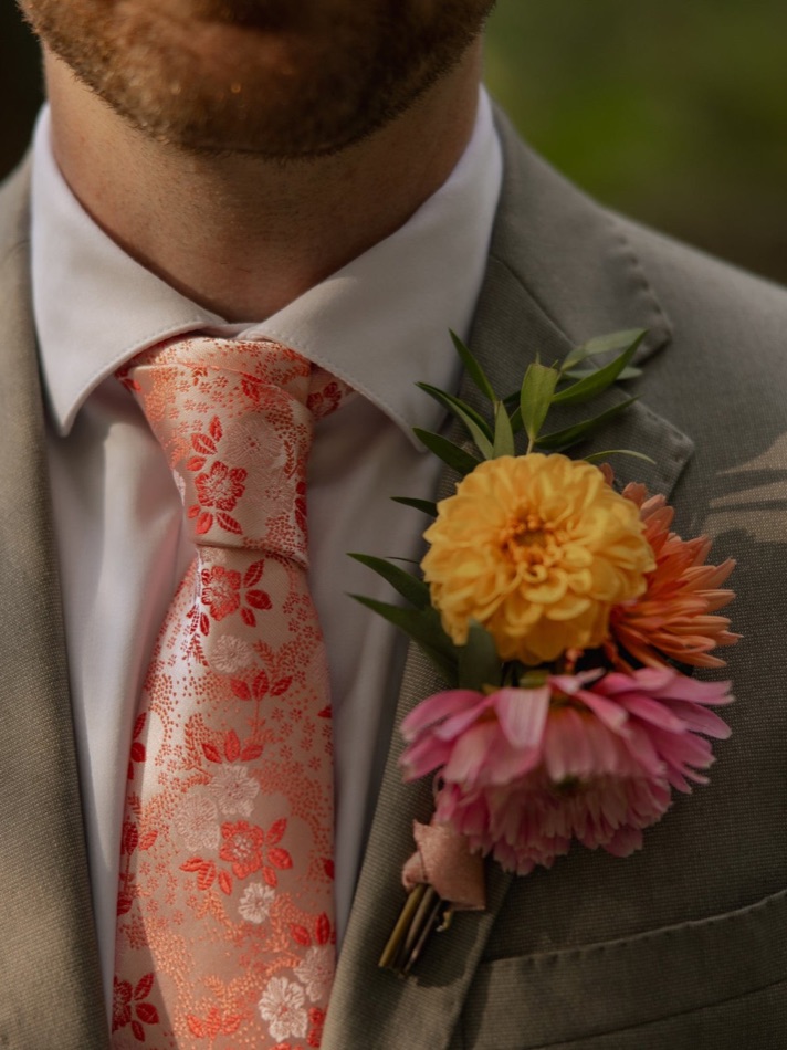 A groom in a sage suit wearing a boutonniere of yellow, coral and pink blooms on his lapel